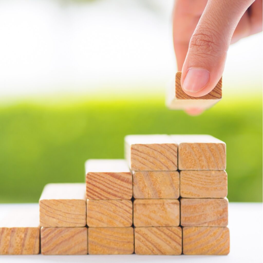 a hand holding a wooden block, adding it to a stacked foundation of wooden blocks.