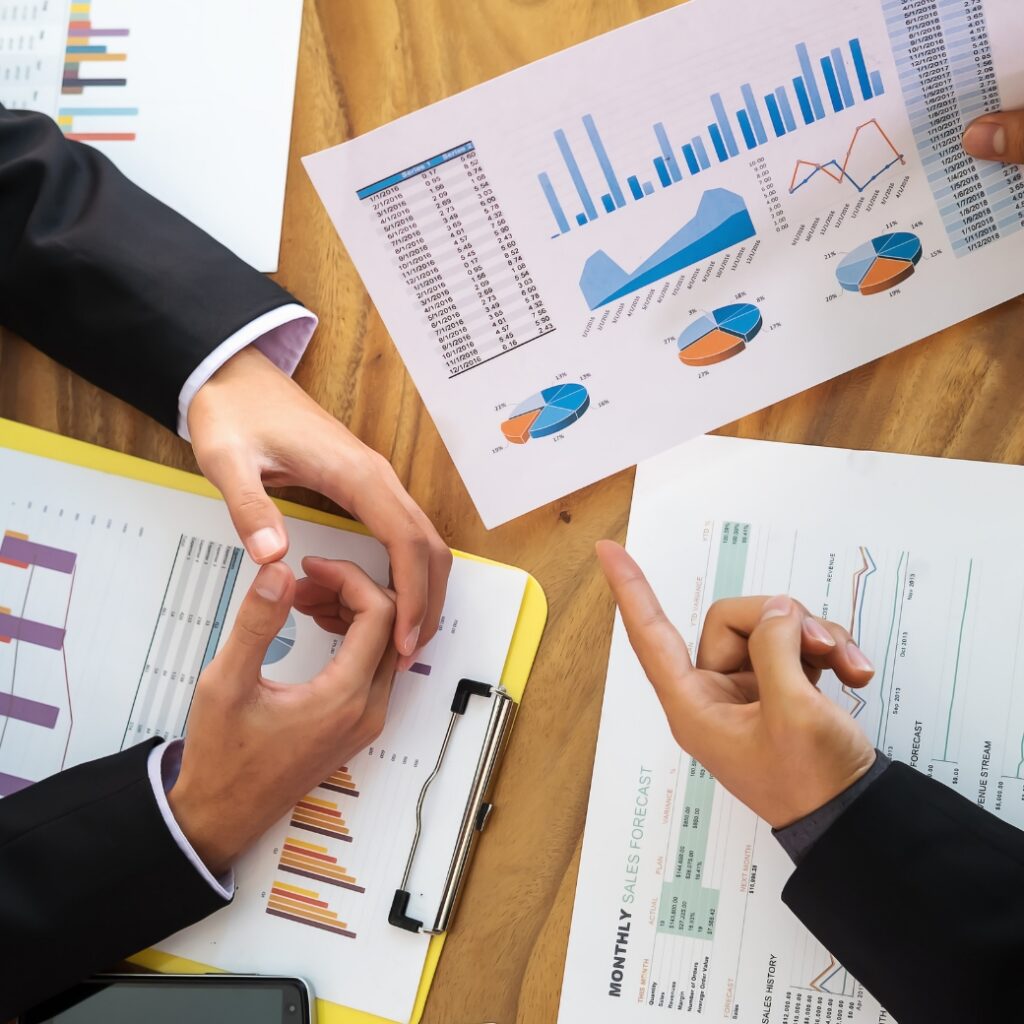 A table with a number of papers scattered with graphs and analytical charts. There are 3 hands visible, analyzing the data.
